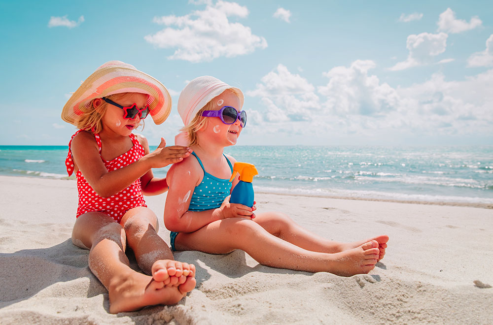 Dve devojke na plaži u kupaćim kostimima, nose naočare za sunčanje i šešire, nanose kremu za sunčanje.