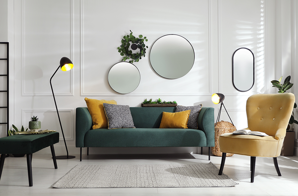 Moderan dnevni boravak u svetlim bojama sa zelenom sofom, žutim jastučićima i okruglim ogledalima na zidu.
