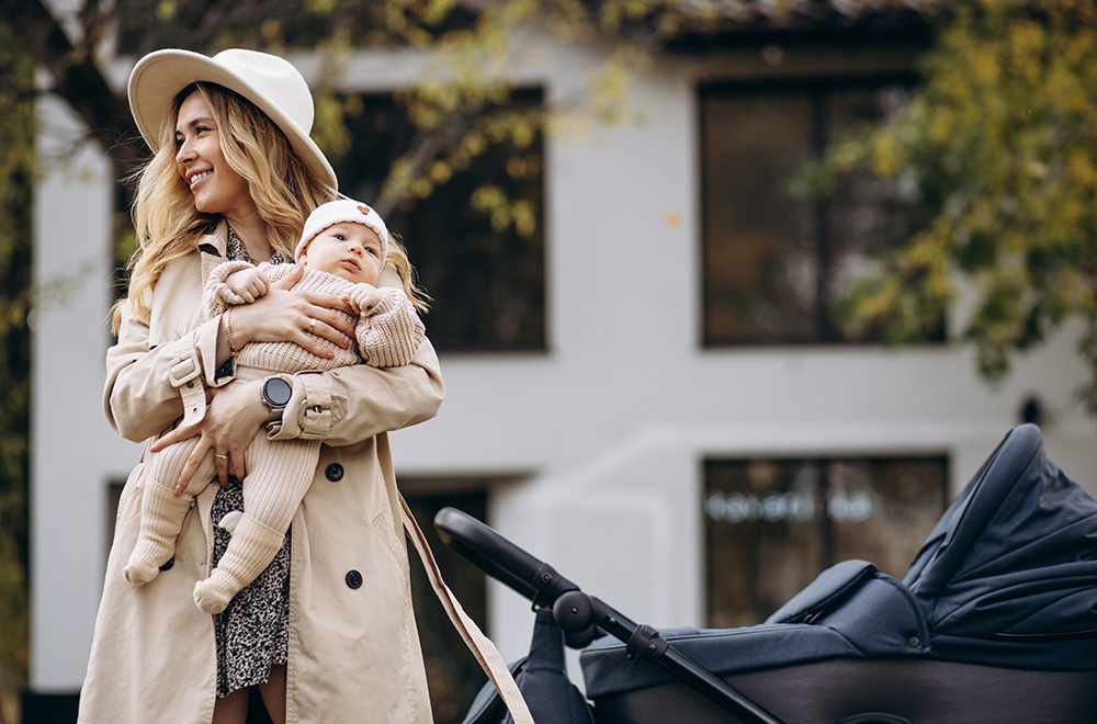 Žena drži bebu u naručju dok šeta pored kolica ispred zgrade